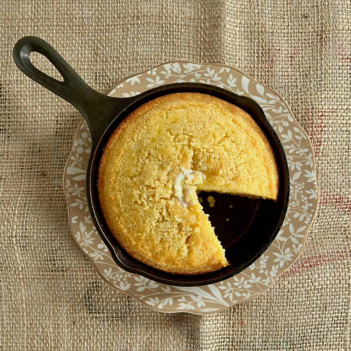 Cut cornbread topped with pat of butter in a cast iron skillet on a brown edged plate from overhead.