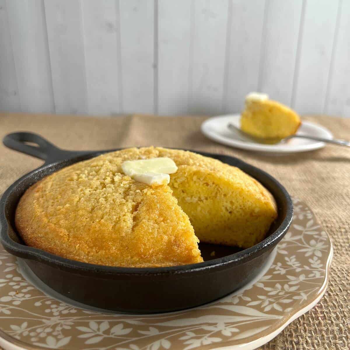 Cut cornbread topped with pat of butter in a cast iron skillet on a brown edged plate with a fork.