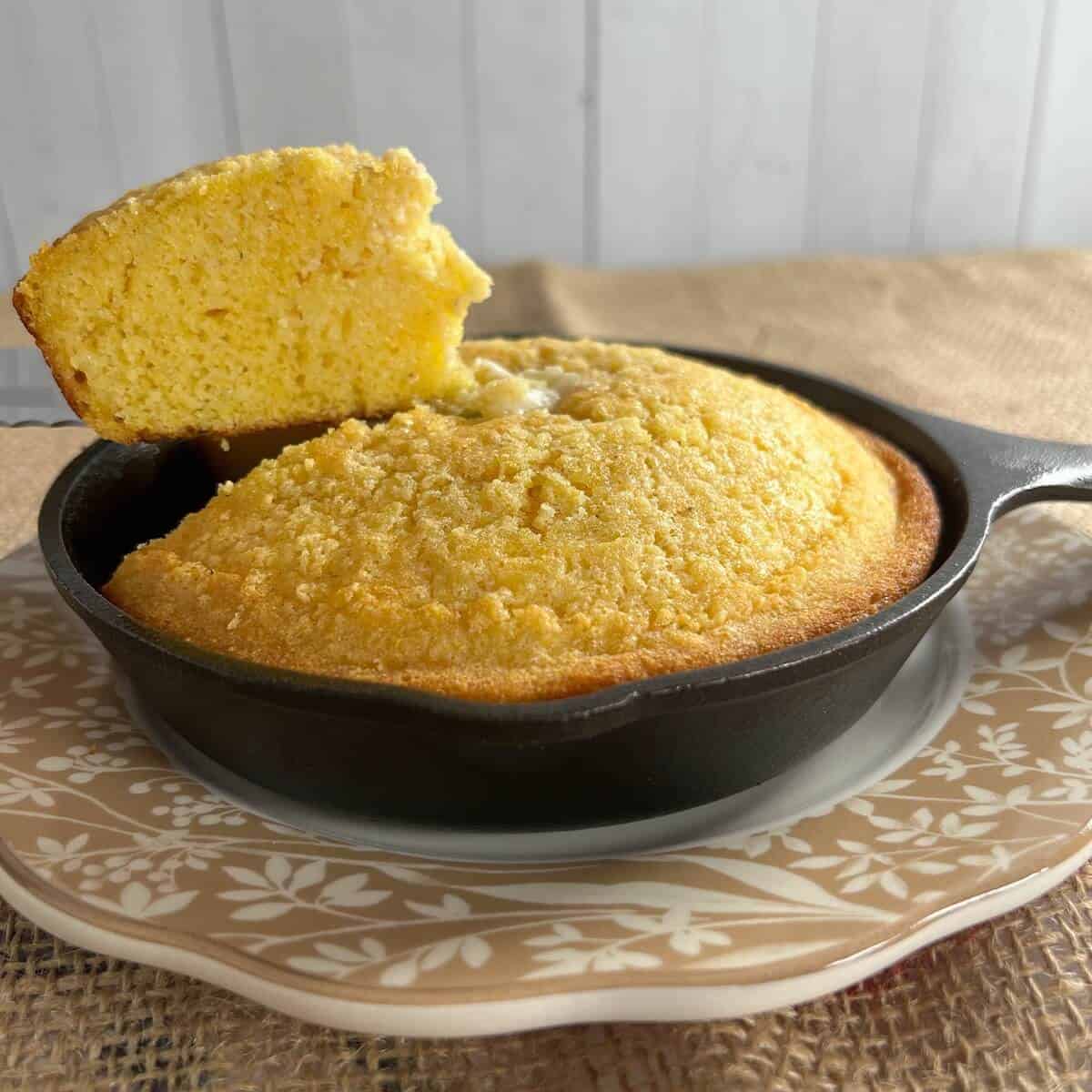Cut cornbread topped with pat of butter in a cast iron skillet on a brown edged plate with a lifted slice.