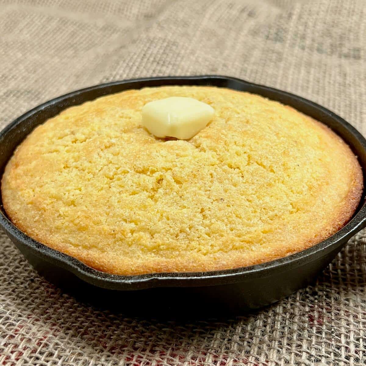 Whole cornbread topped with pat of butter in a cast iron skillet on a piece of burlap.
