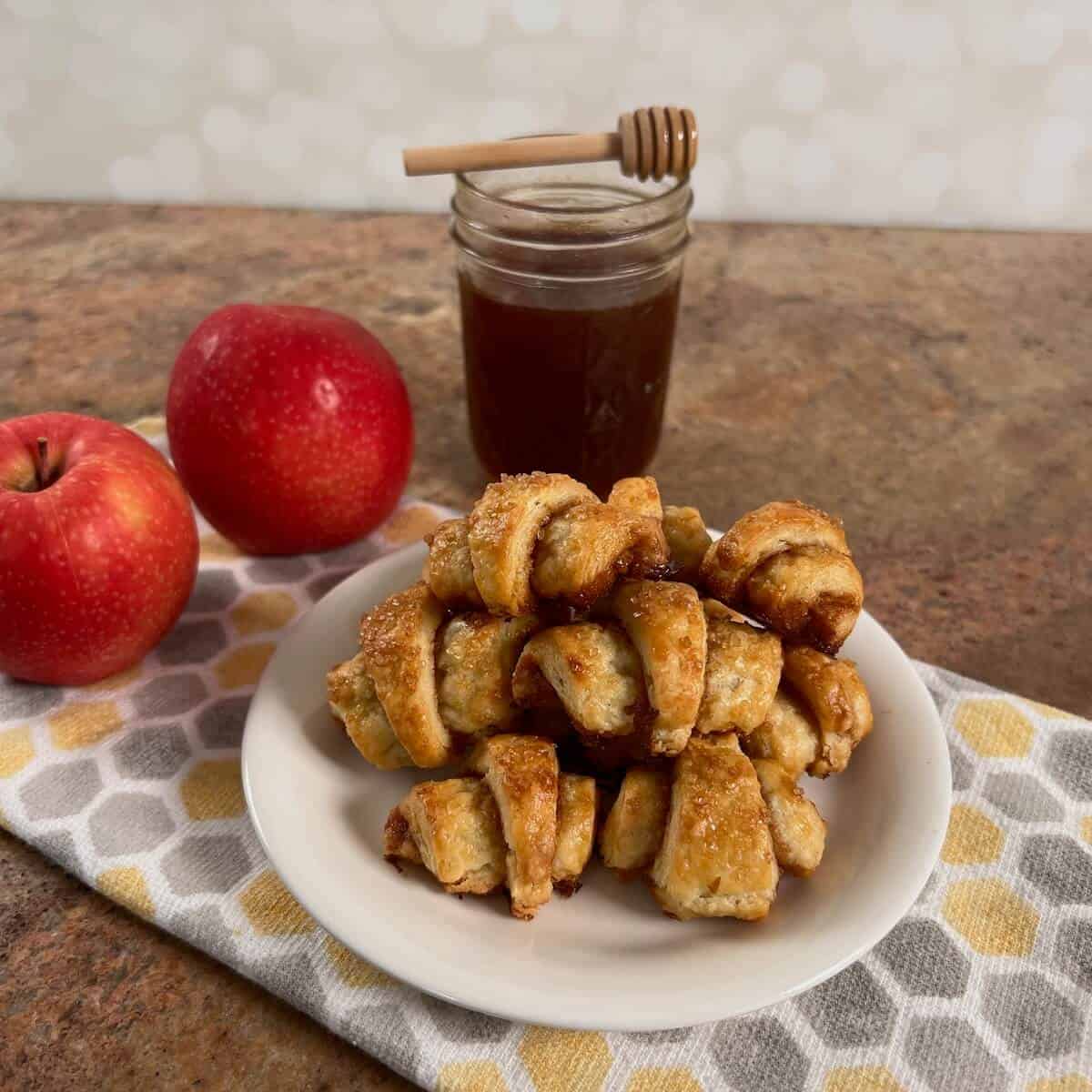 Apple Cinnamon Rugelach stacked on a white plate in front of 2 apples and a jar of boiled cider.