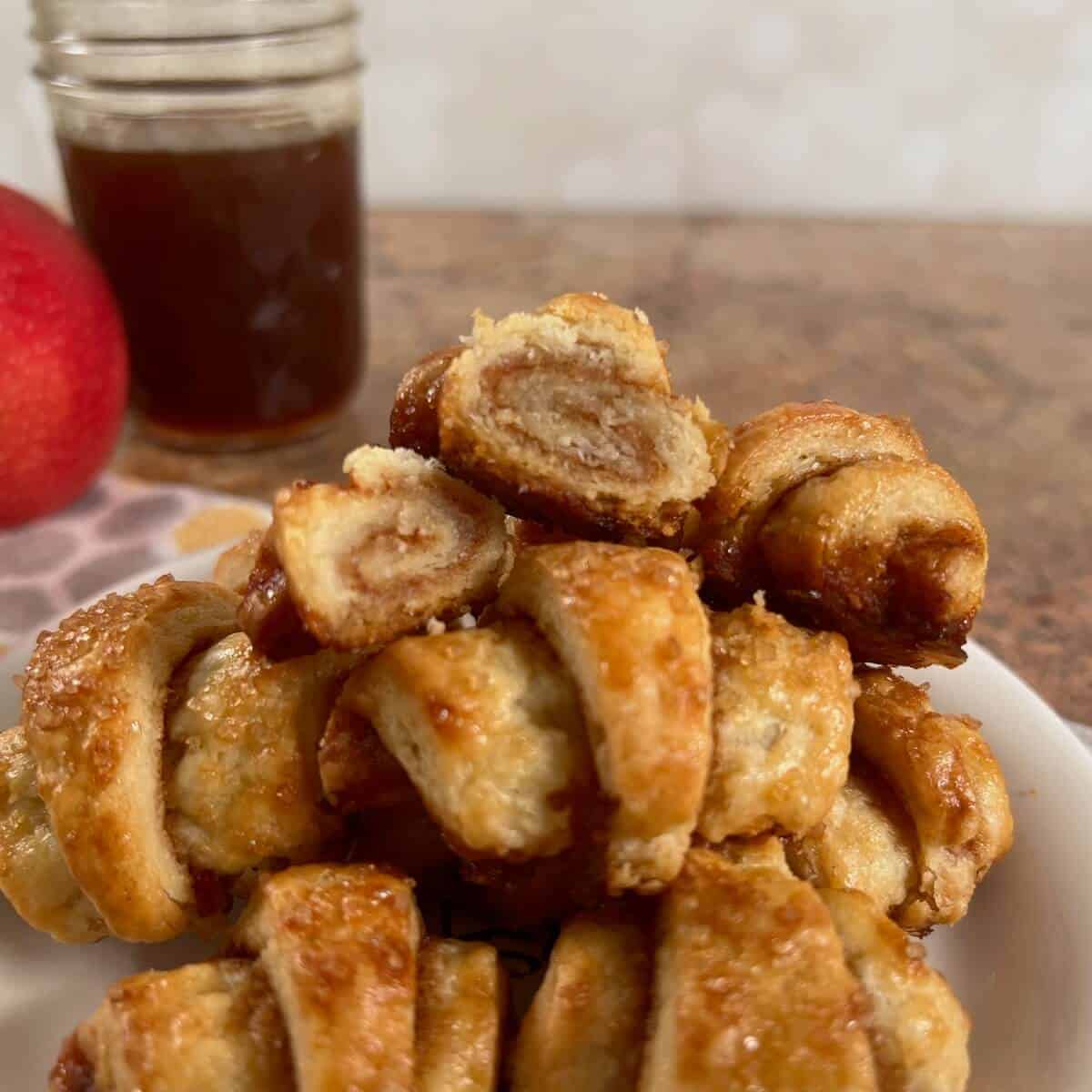 Apple Cinnamon Rugelach stacked on a white plate, with the top one cut in half, in front of an apple and a jar of boiled cider.