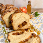 Sliced apple honey babka on a plate with an apple and honey in a jar and bowl behind all on a honeycomb-decorated towel.