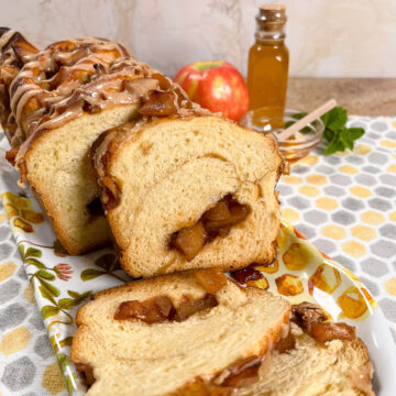 Sliced apple honey babka on a plate with an apple and honey in a jar and bowl behind all on a honeycomb-decorated towel.