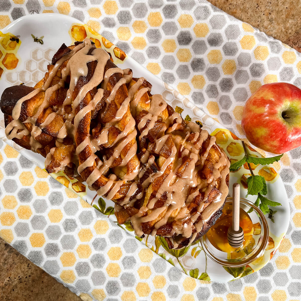 Whole apple honey babka on a plate with a bowl of honey and an apple all on a honeycomb-decorated towel.