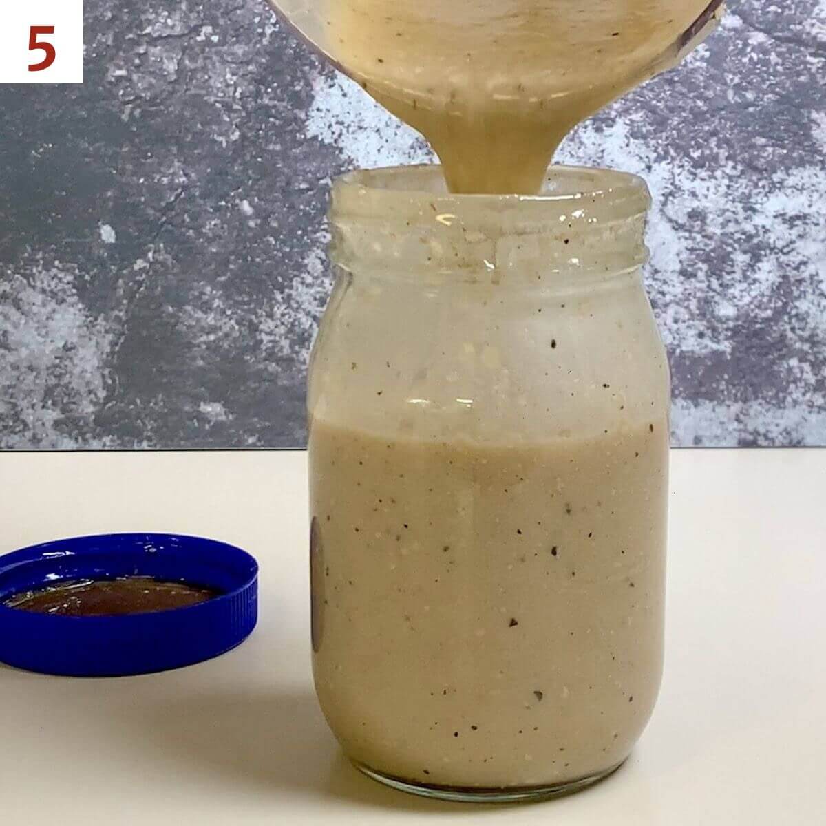 Pouring Caesar salad dressing into a glass jar on a counter.