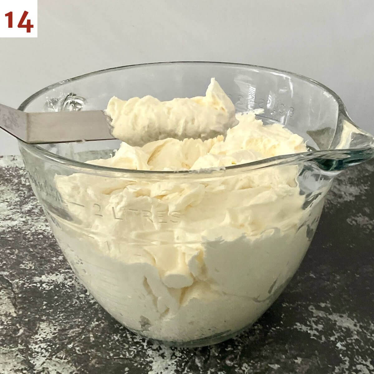 Vanilla buttercream frosting in a large glass measuring bowl with some on a lifted spatula.