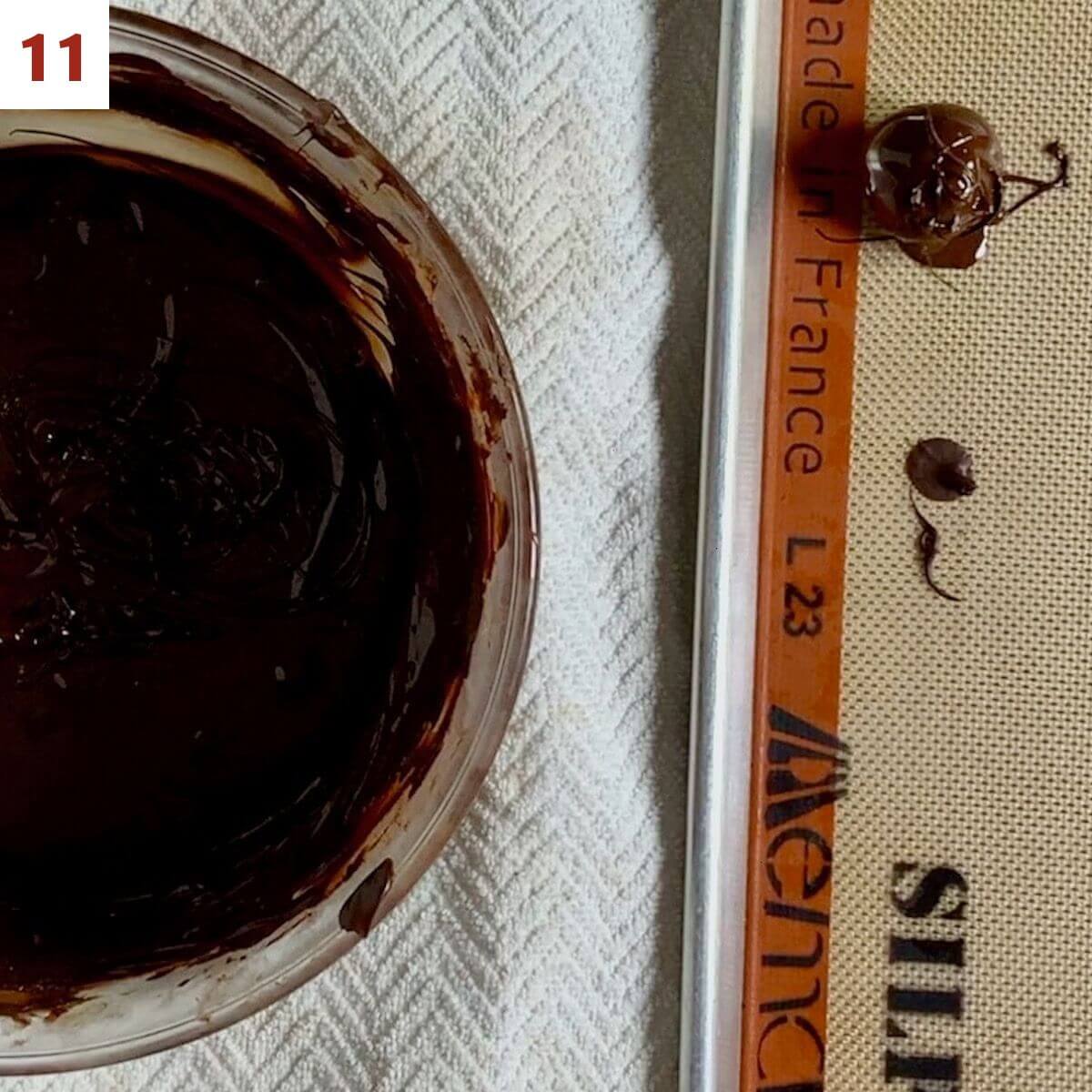 Single dipped chocolate truffle drying on baking pan lined with a Silpat silicone mat next to bowl of tempered chocolate.