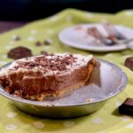 Half chocolate cream pie in the pan with 2 forks on a plate in background on a green polka dot towel.