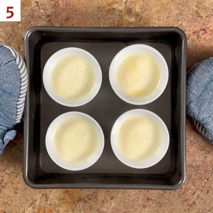 Four custard base-filled ramekins in a pan filled with hot water.