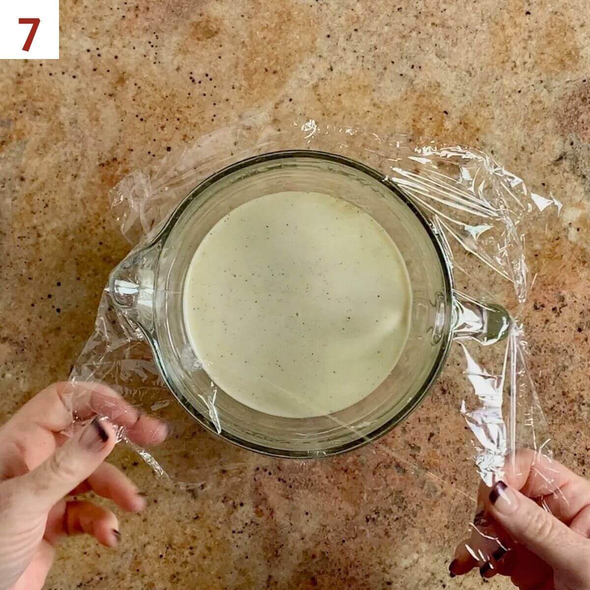 Covering eggnog in a glass pitcher with plastic wrap.