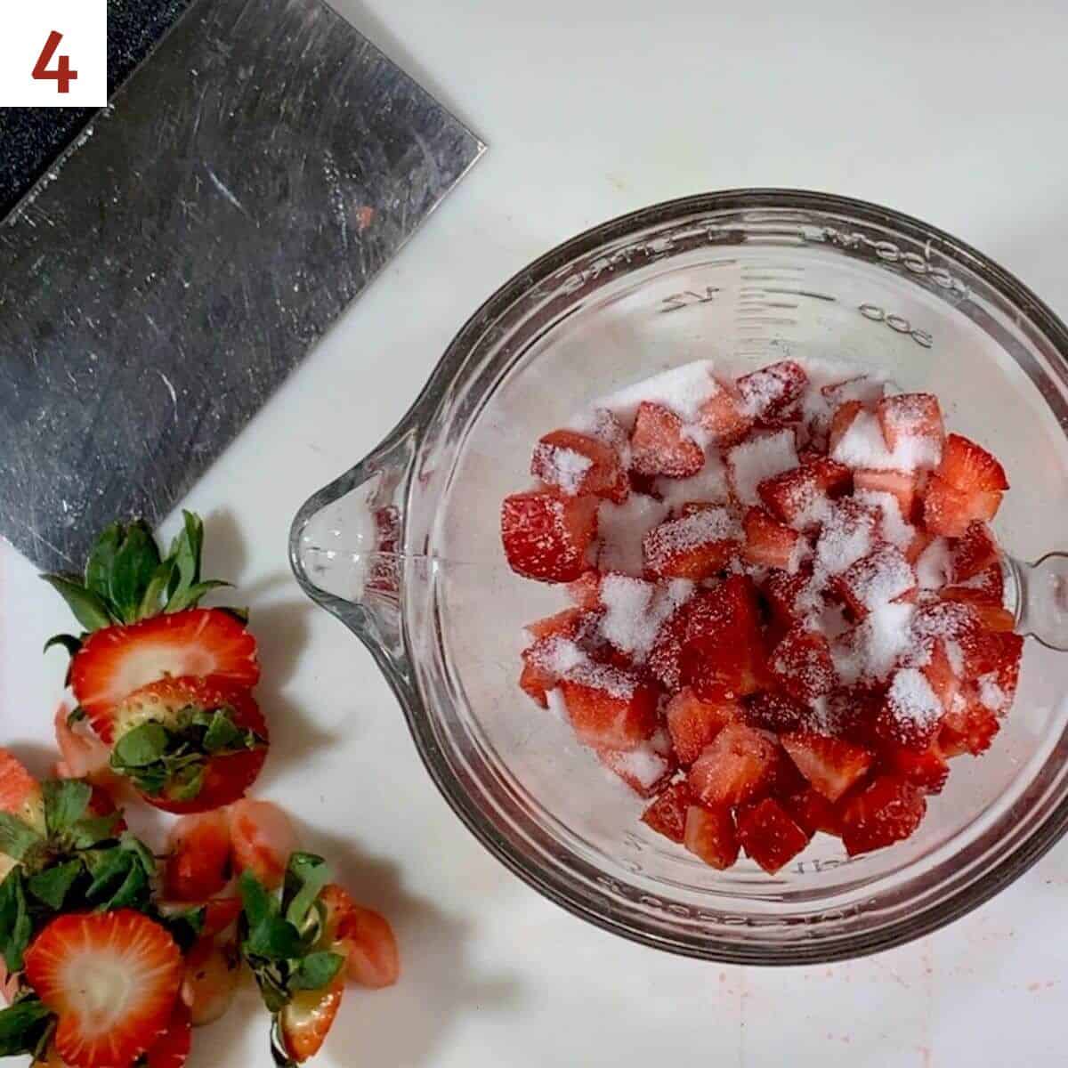 Chopped strawberries in a glass bowl sprinkled with sugar with strawberry tops and bench scraper on the side.