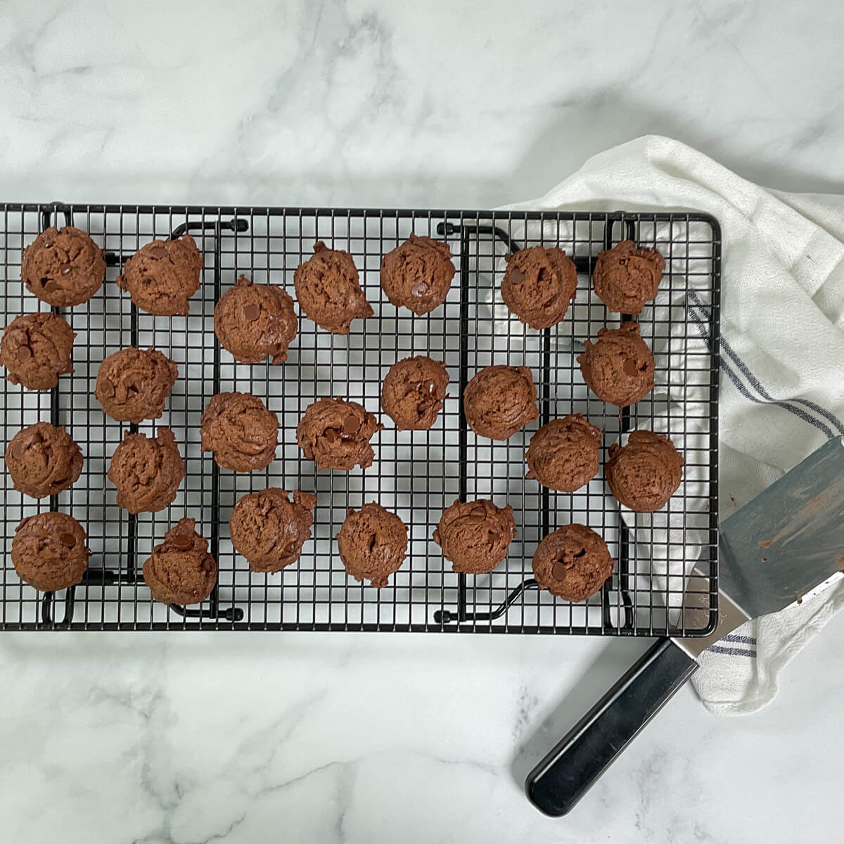 Baked cookies on a black cooling rack next to a large spatula and a white towel with a blue stripe from overhead.