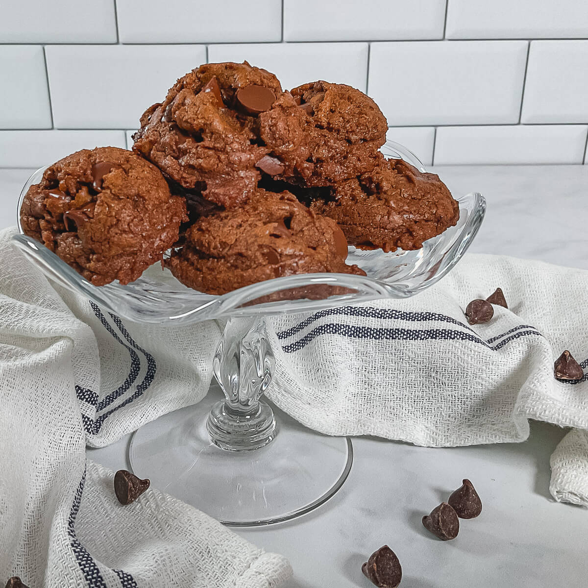 Mocha Chocolate Truffle Cookies stacked on a glass cake stand surrounded by a white towel with a blue stripe & chocolate chips.