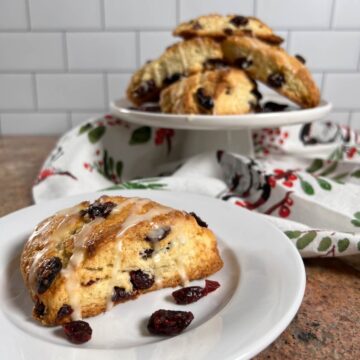 Orange Cranberry scone on a white plate in foreground & stacked on a white cake stand in background.