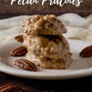 Pecan pralines stacked on a white plate with pecans & ivory scarf behind Pinterest banner.