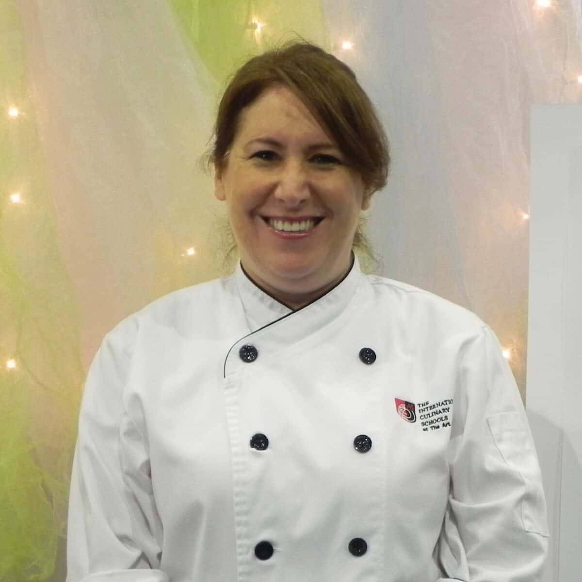 Tammy dressed in her culinary school chef's coat in front of a pastel background lit with fairy lights.