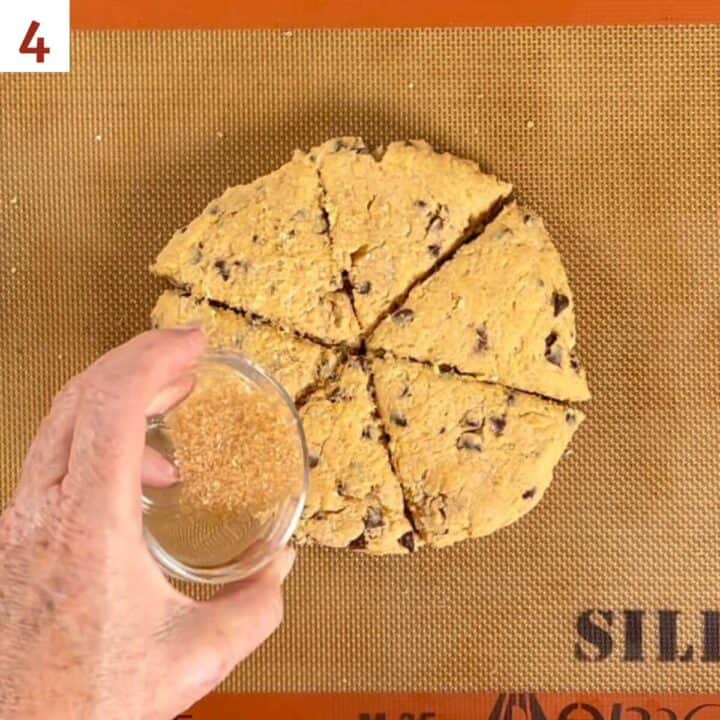 Scone dough shaped into a 6-inch disk on a baking pan, cut into 6 portions, and being sprinkled with coarse sugar.