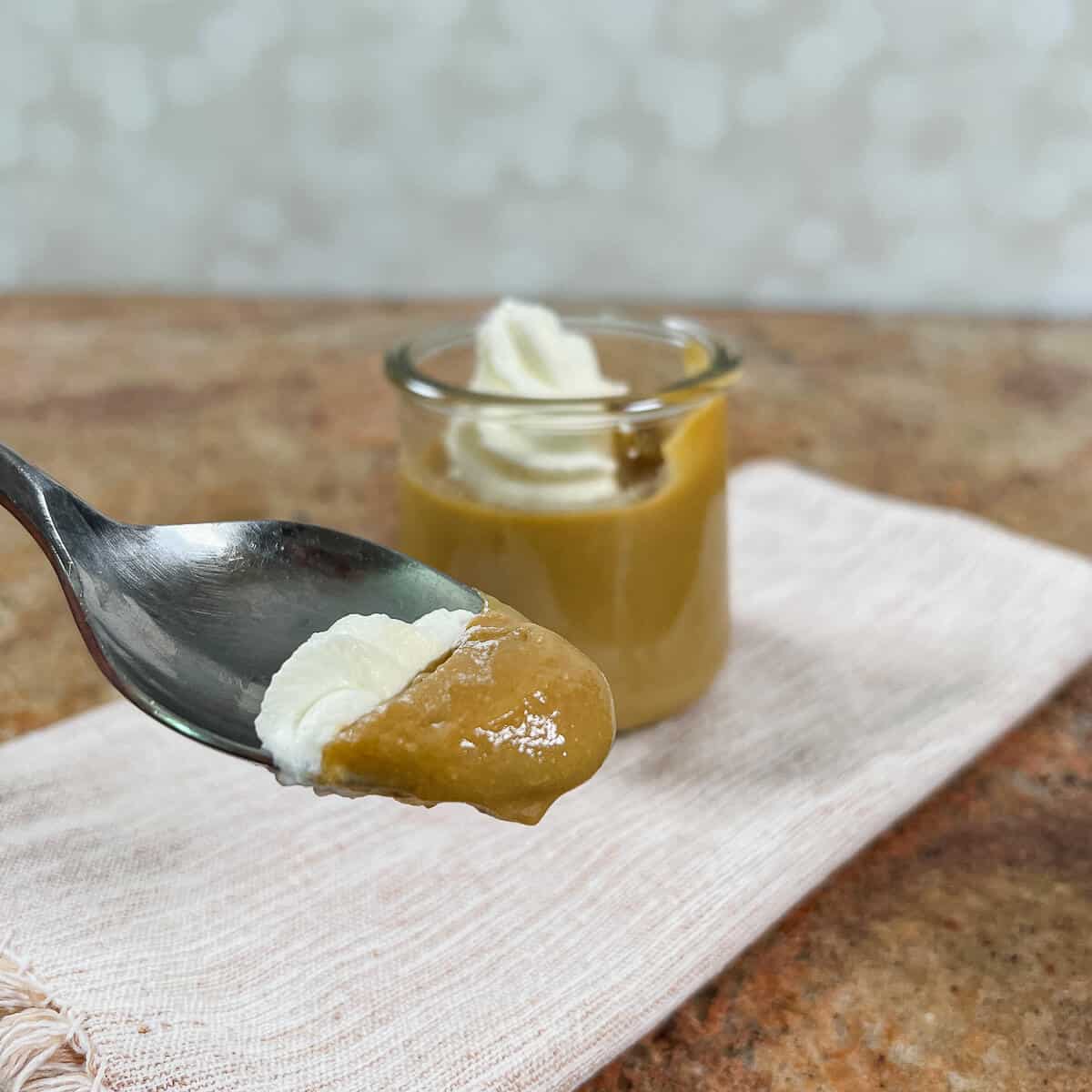 Spoonful of butterscotch pudding and whipped cream in front of the dish of pudding on a light brown napkin.