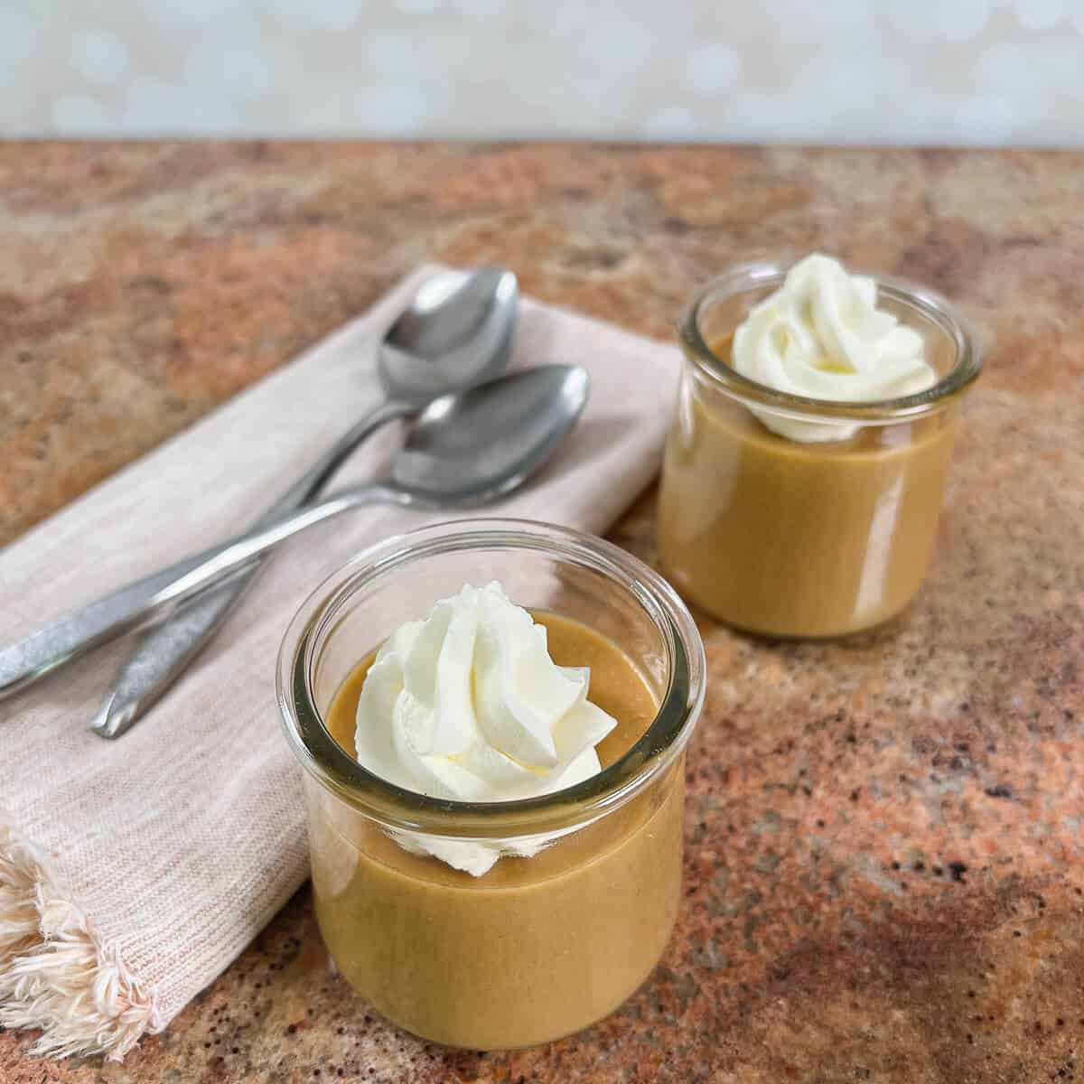 Two servings of butterscotch pudding with whipped cream in glass dishes next to two spoons on a light brown napkin.