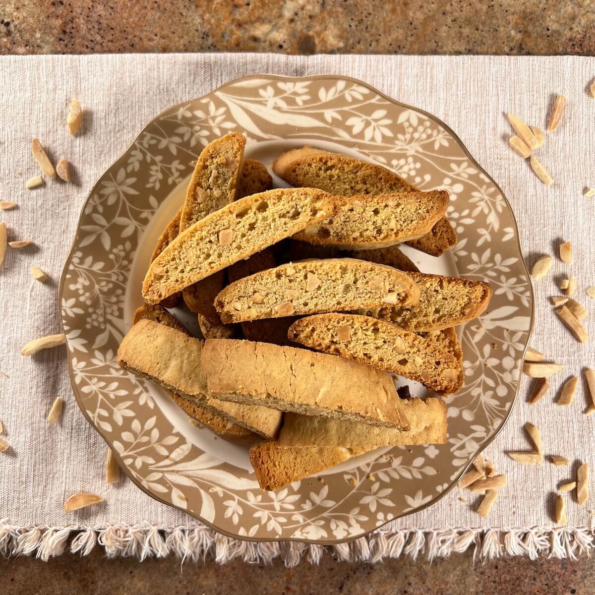 Mandel bread piled onto a white plate with a brown flowered edging surrounded by slivered almonds on a tan placemat from overhead.