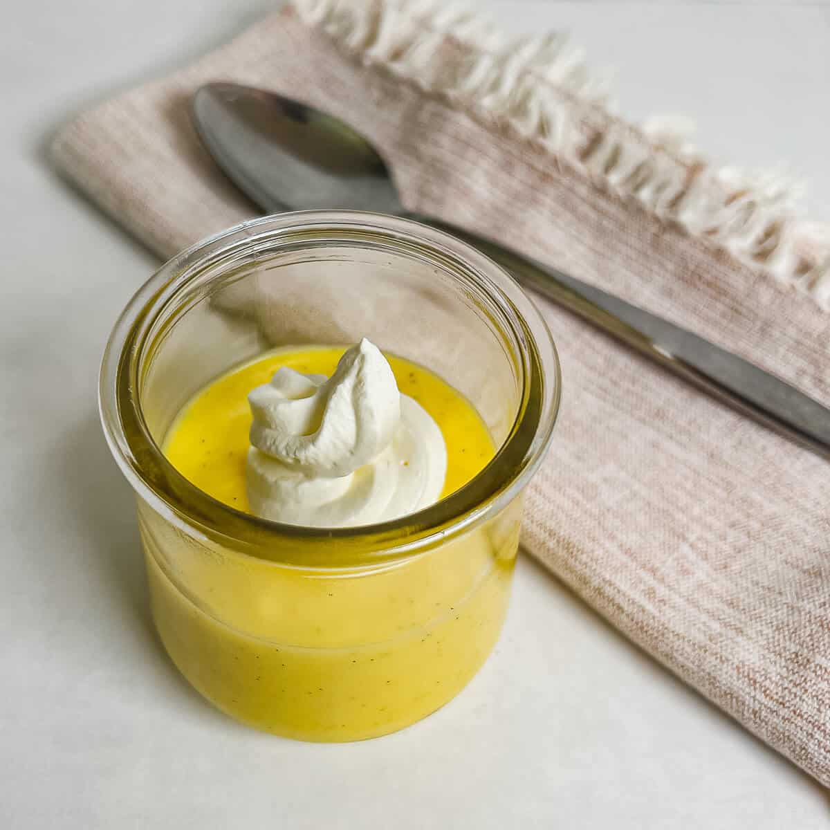 Pastry cream in a glass dish topped with whipped cream with a spoon on a towel behind.