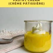 Pastry cream in a glass dish topped with whipped cream with a spoon on a towel behind Pinterest banner.