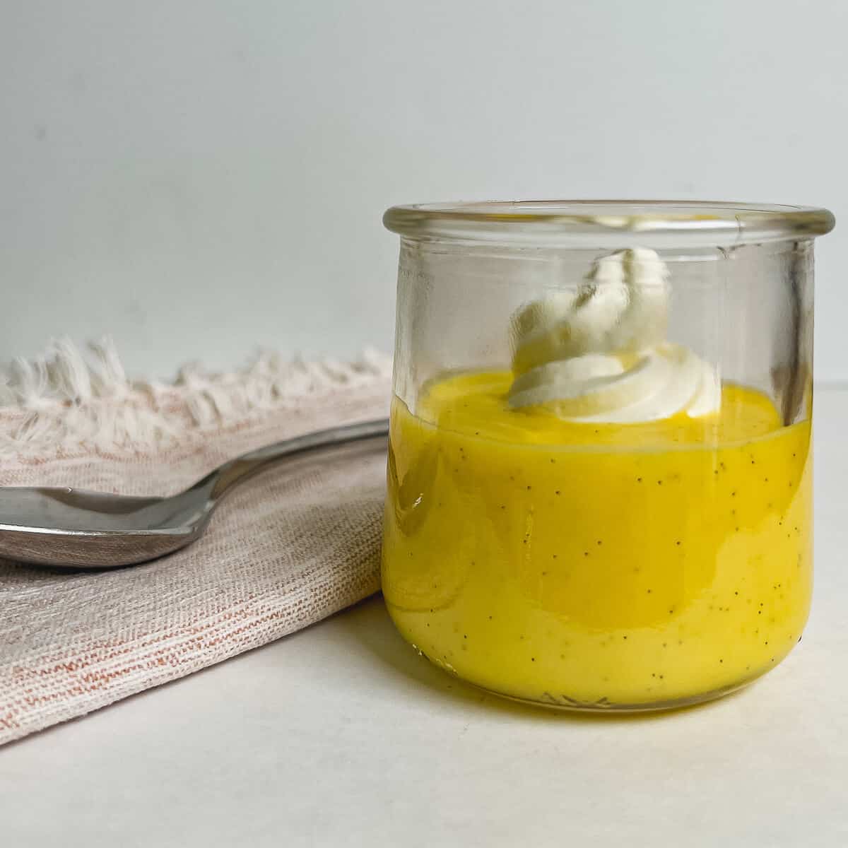 Pastry cream in a glass dish topped with whipped cream with a spoon on a towel behind.