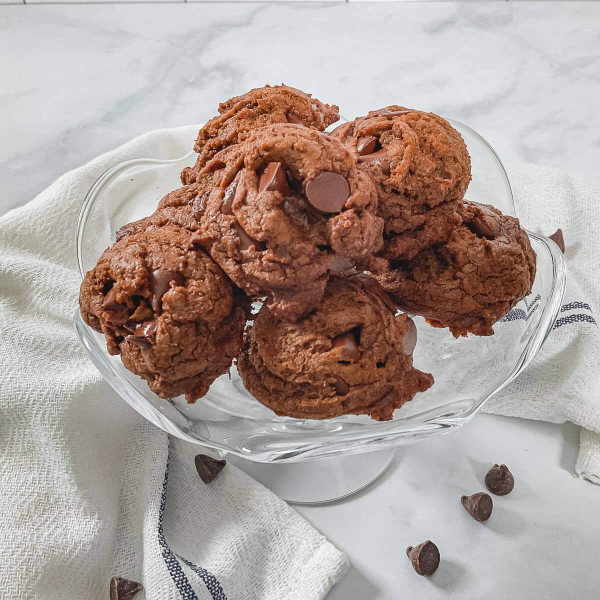 Mocha Chocolate Truffle Cookies stacked on a glass cake stand surrounded by a white towel with a blue stripe & chocolate chips from overhead.