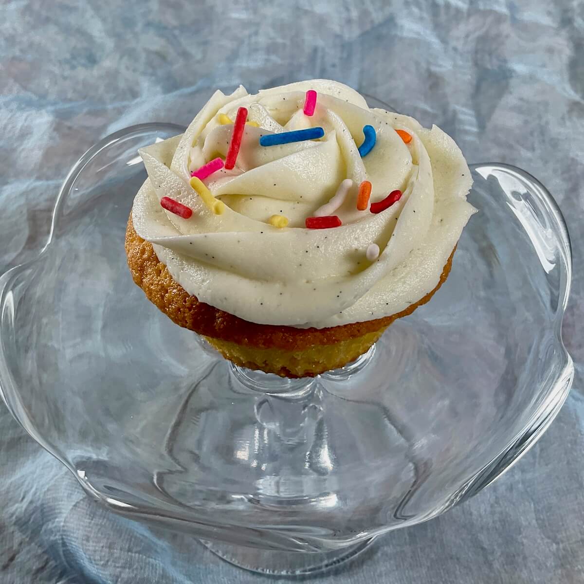 Vanilla cupcake frosted with vanilla buttercream topped with sprinkles on a glass cake stand.