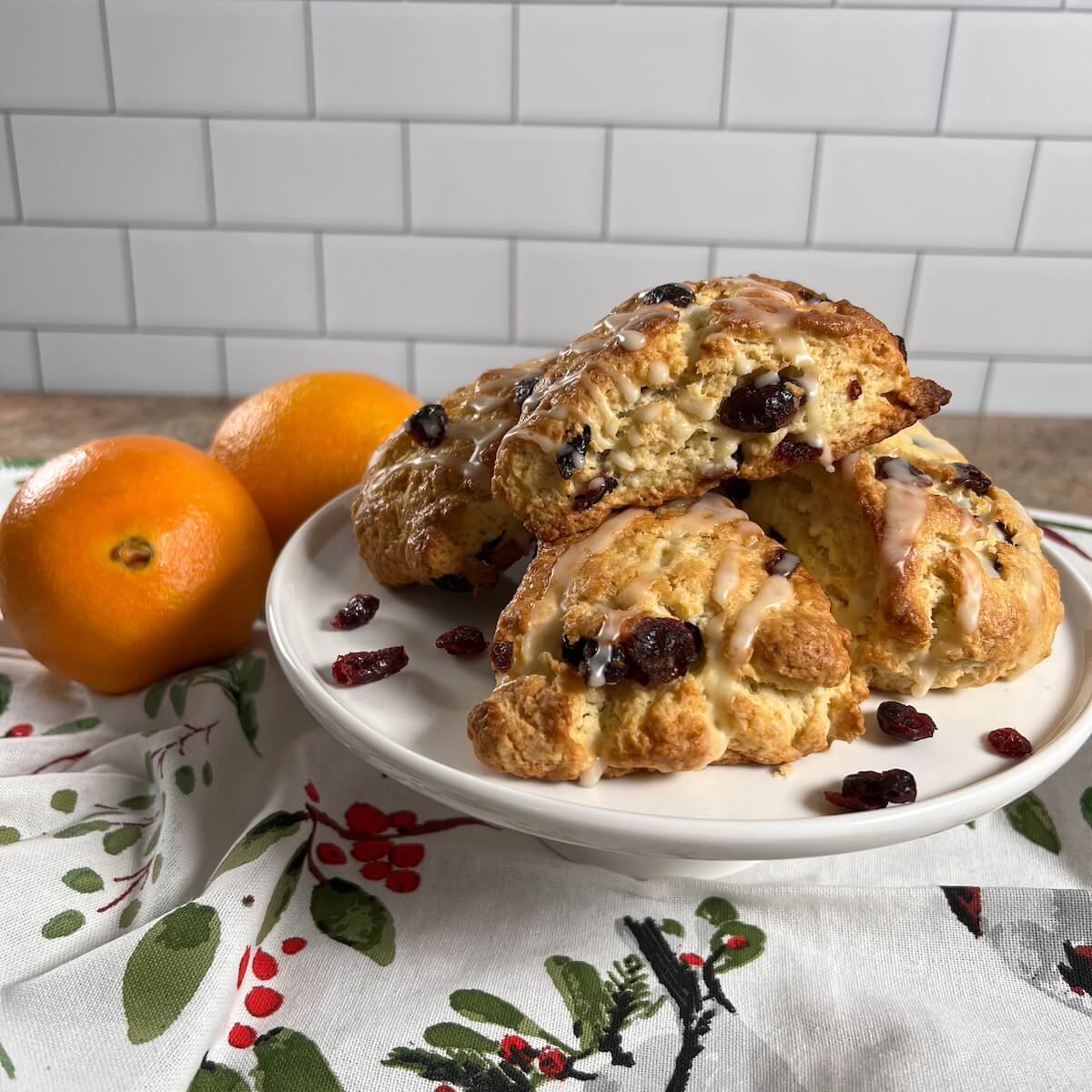 Stack of Orange Cranberry Scones on a white cake stand with scattered dried cranberries and next to 2 oranges.