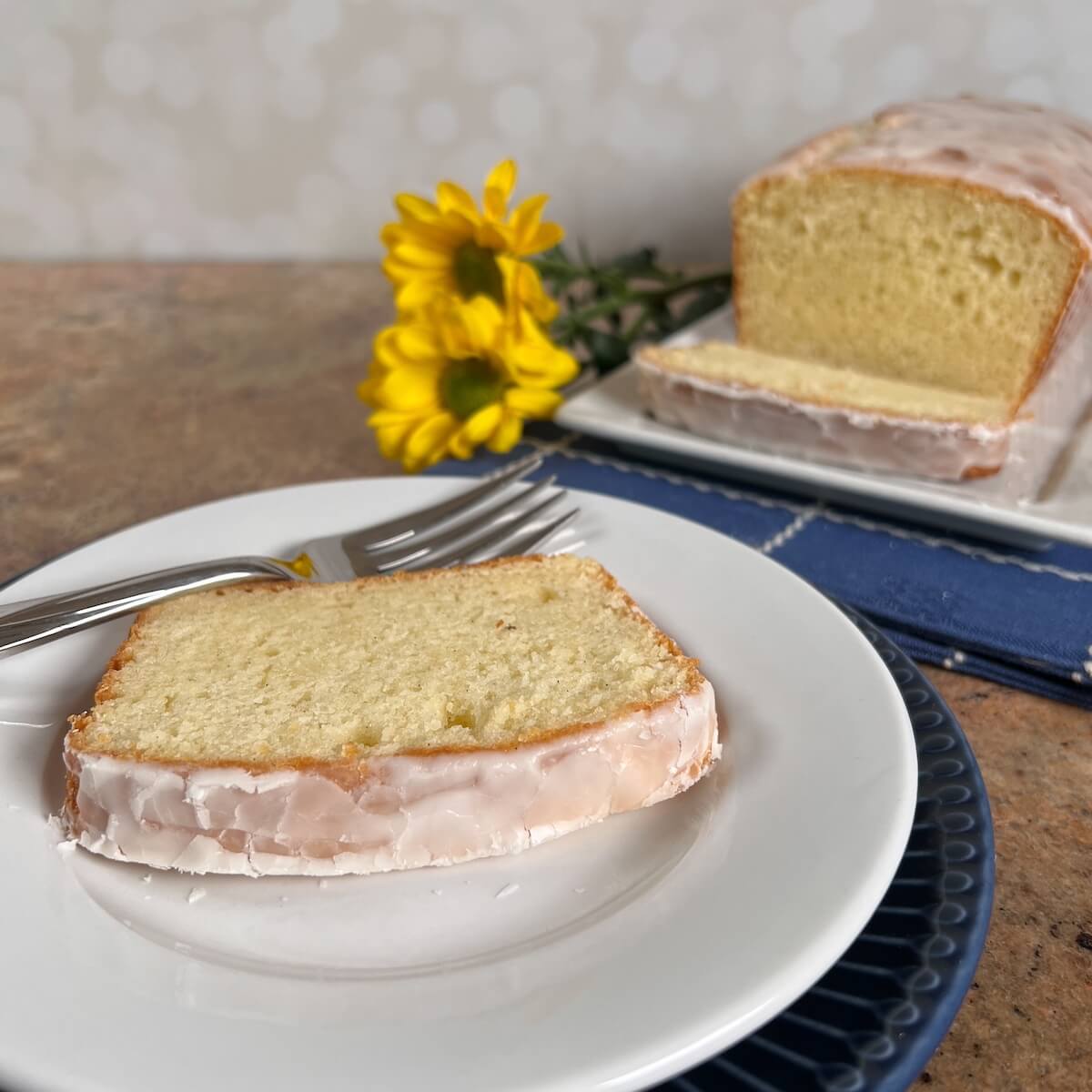 Slice of Limoncello Cake on a white plate with a fork next to the sliced loaf sliced on a white plate with yellow flowers.