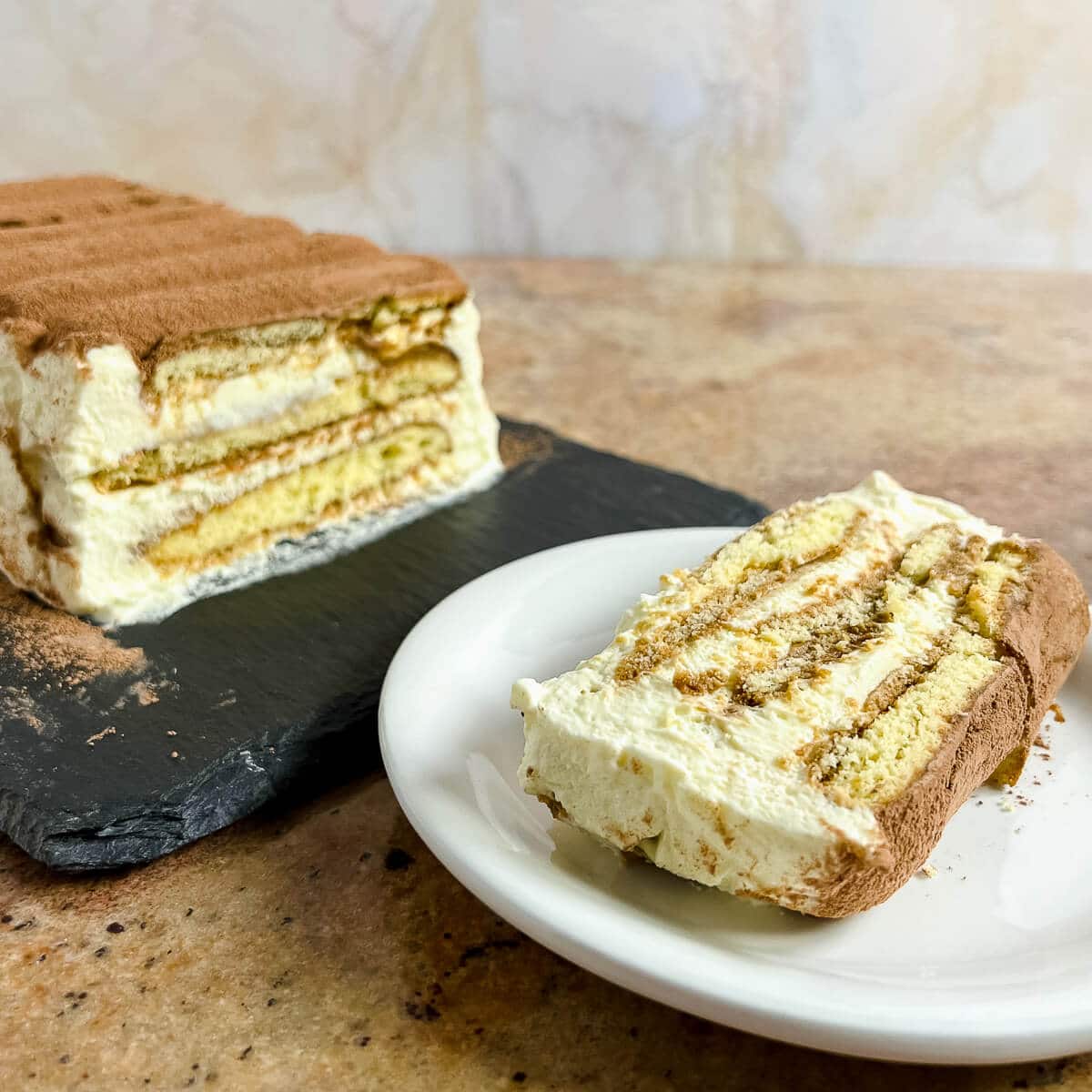 Slice of Tiramisu loaf on a plate next to the loaf on a black platter.