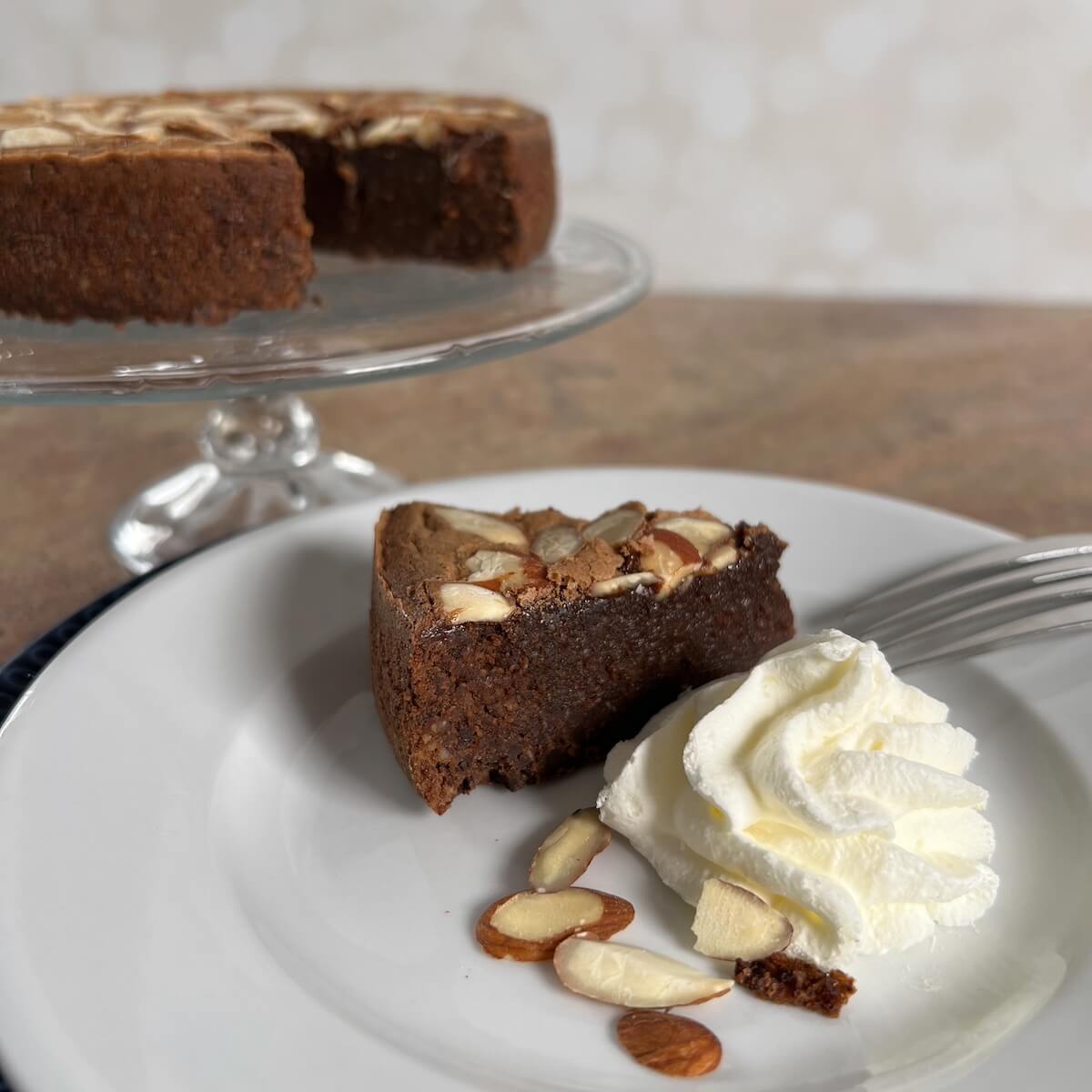 Slice of Flourless Chocolate Almond Cake next to piped whipped cream with the cake behind on a cake stand.