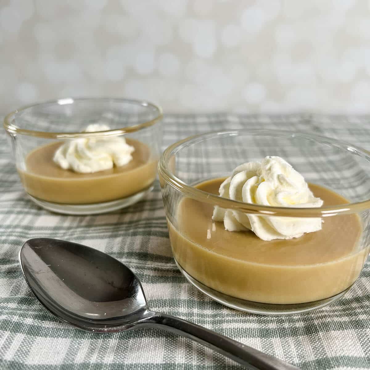 Two servings of maple pudding each with a dollop of whipped cream next to a spoon on a green checked cloth.