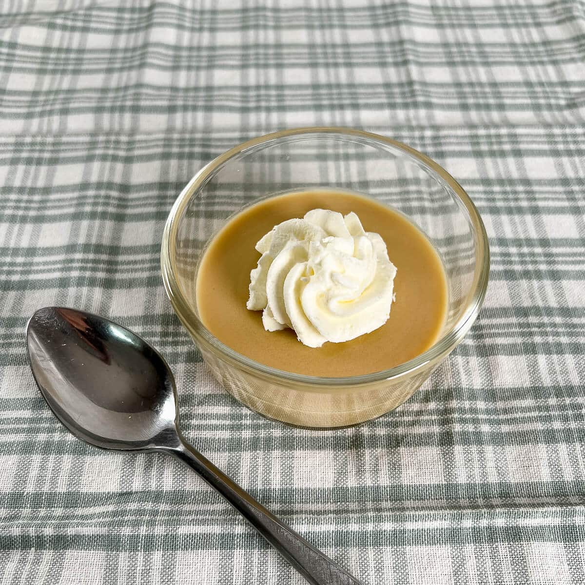 A serving of Maple pudding with a dollop of whipped cream next to a spoon on a green checked cloth.