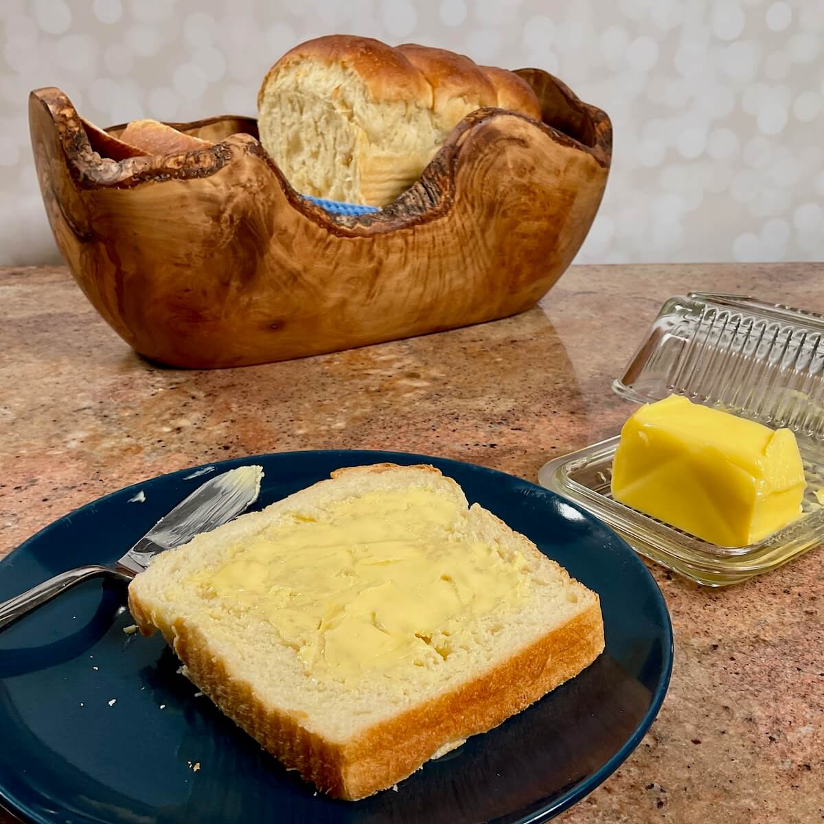 Buttered slice of sourdough discard sandwich bread on a blue plate next to butter with sliced loaf in a wooden bowl behind it.