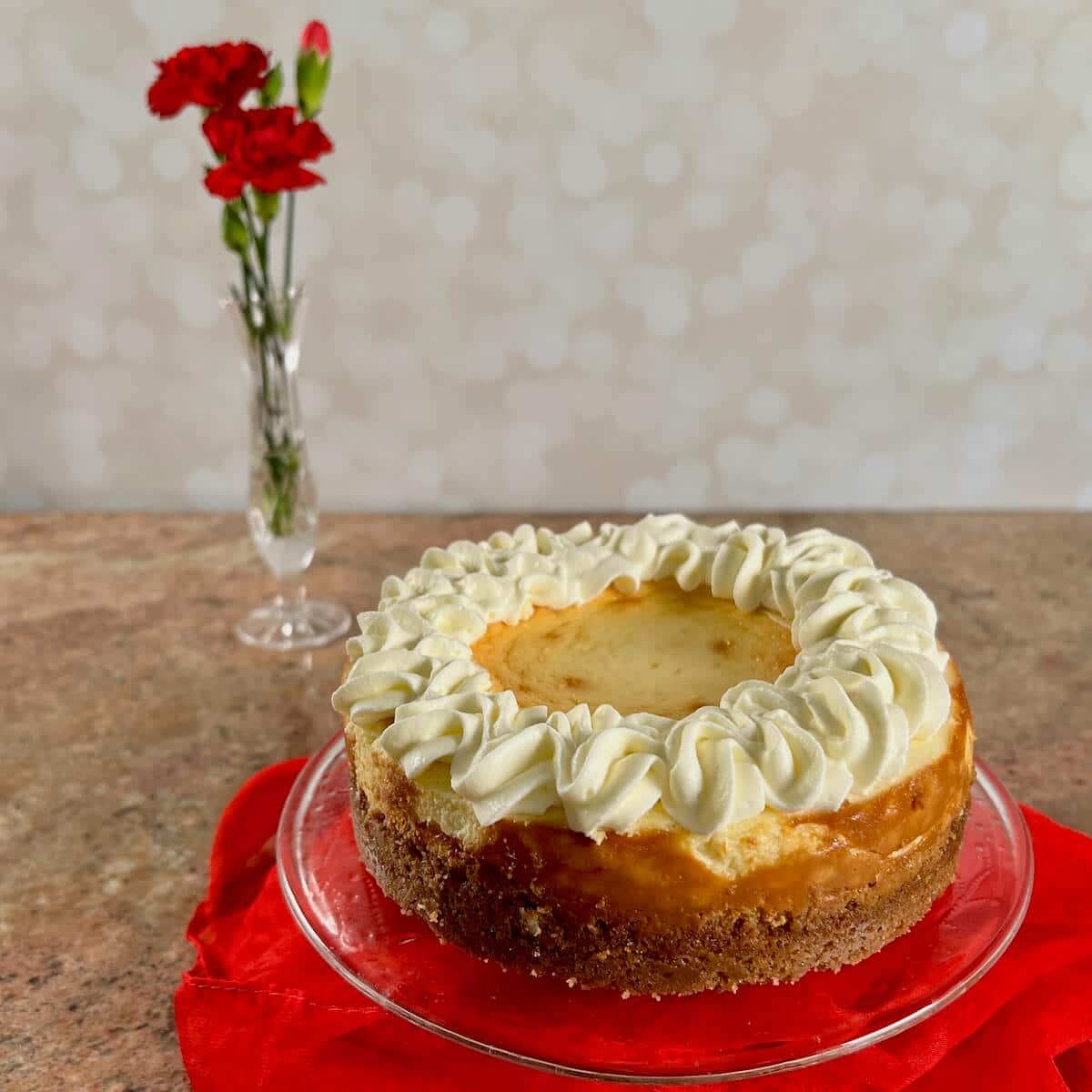 Whole white wine cheesecake on a cake stand wrapped in a red scarf next to a vase of red carnations.