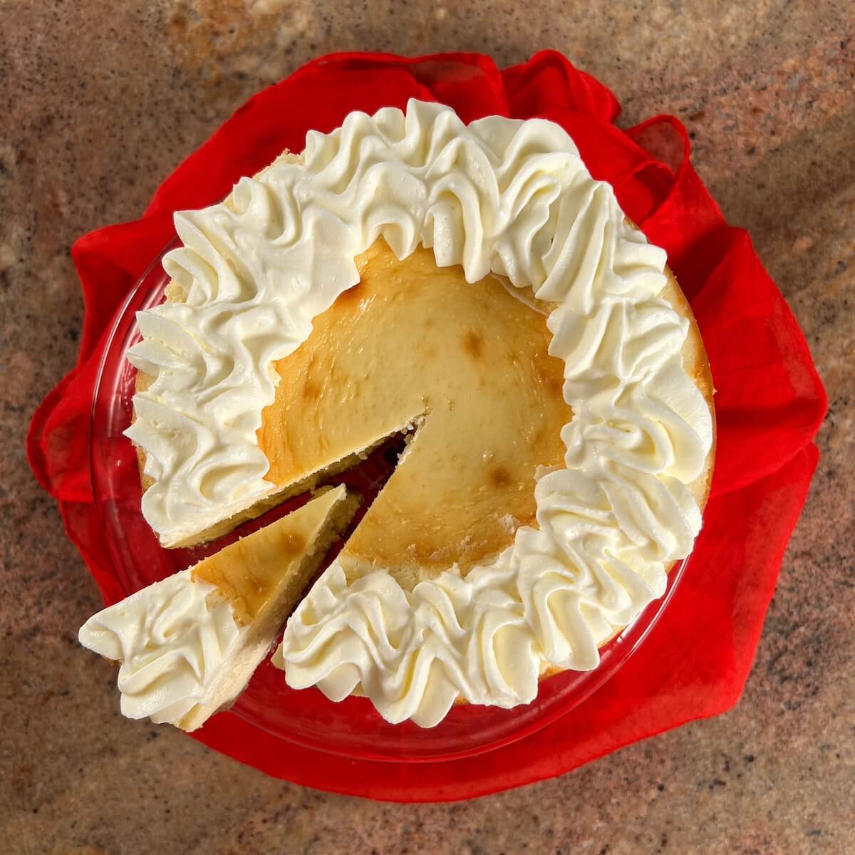 White wine cheesecake with a slice pulled out on a cake stand wrapped in a red scarf from overhead.