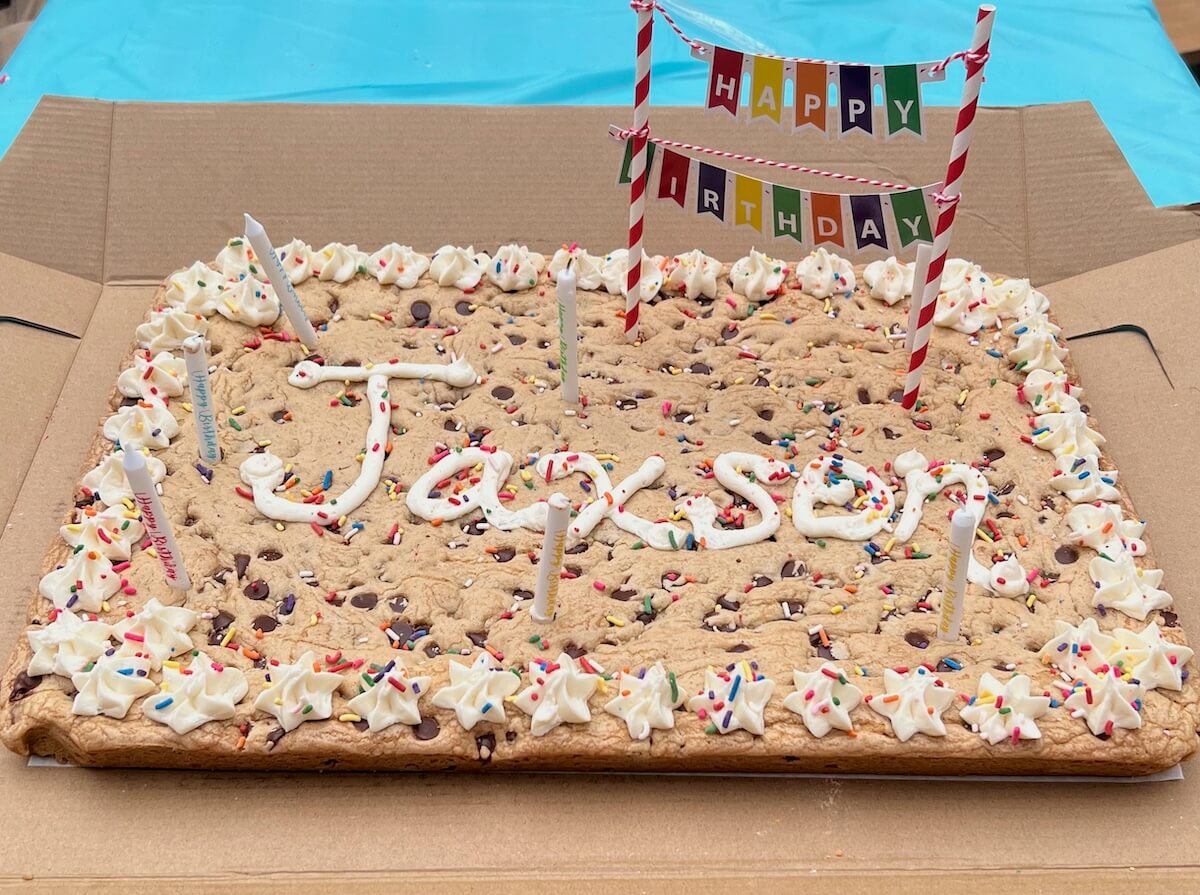 Giant rectangular sheet cookie cake decorated with sprinkles and frosting with a happy birthday banner and candles.