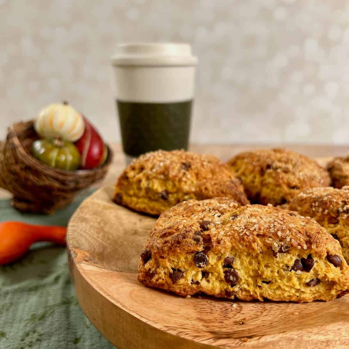 Pumpkin Chocolate Chip Scones stacked on a wooden cake stand in front of a coffee cup and basket of small pumpkins.