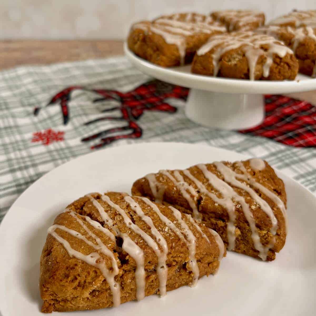 Two gingerbread scones on a white plate in front of gingerbread scones sitting on a white cake stand on a green plaid towel.