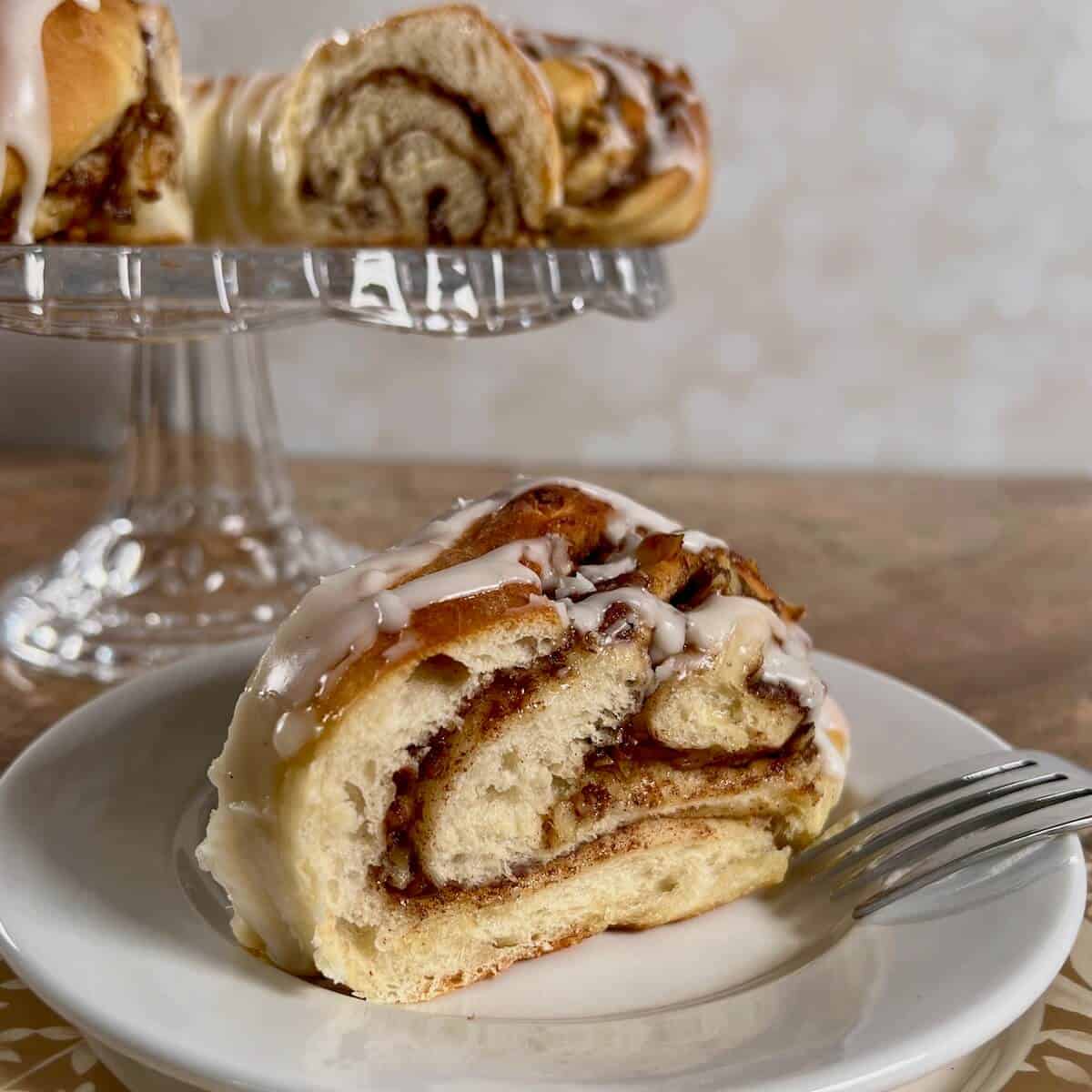 Slice of Swedish Tea Ring on a white plate with a fork in front of the rest of the ring on a glass cake stand.