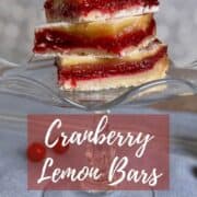 Cranberry Lemon Bars stacked on a glass cake stand with fresh cranberries and sliced lemon below on a blue napkin with white stripes Pinterest banner.