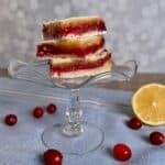 Cranberry Lemon Bars stacked on a glass cake stand with fresh cranberries and sliced lemon below on a blue napkin with white stripes.
