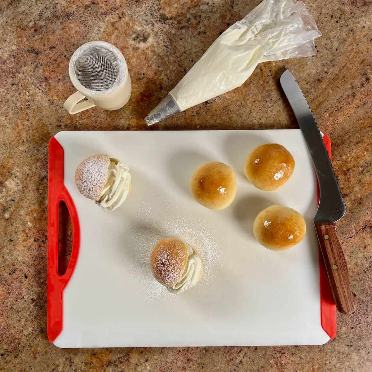 Two finished Scottish Cream Buns on a cutting board beside three unfilled cream buns, a knife, a piping bag with whipped cream, and a powdered sugar duster.