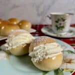 Two finished Scottish Cream Buns on a china plate in front of a china cup and saucer and a plate of unfilled cream buns.