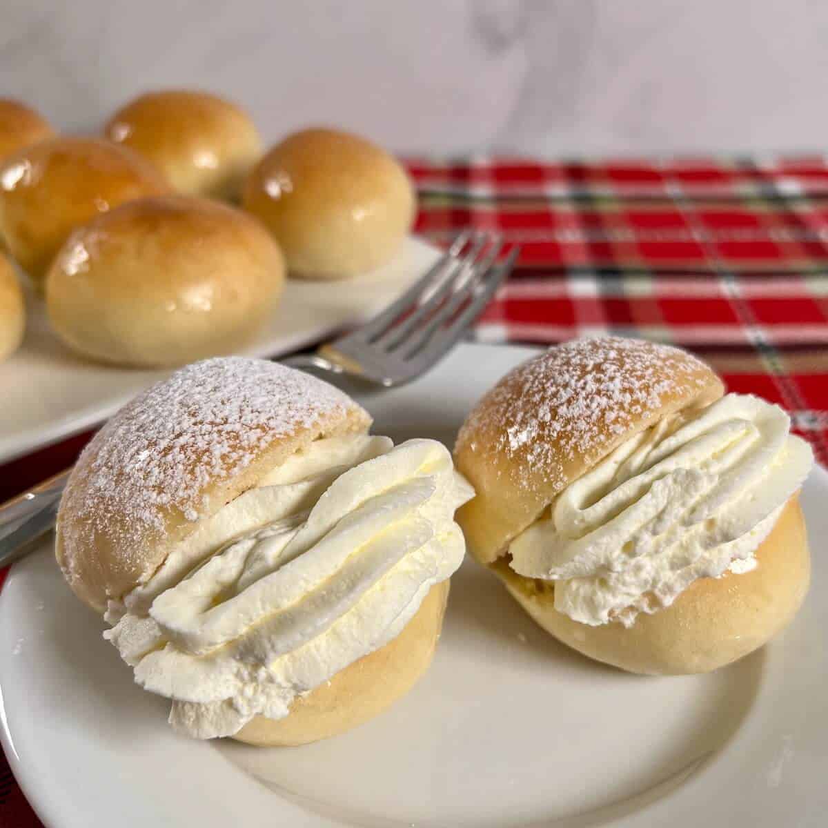 Two finished Scottish Cream Buns on a white plate with a fork beside a plate of unfilled cream buns all on a red plaid napkin.