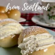 Two finished Scottish Cream Buns on a chine plate in front of a china cup and saucer and a plate of unfilled cream buns Pinterest banner.