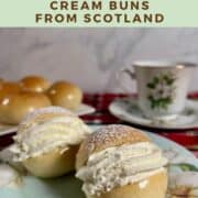 Two finished Scottish Cream Buns on a chine plate in front of a china cup and saucer and a plate of unfilled cream buns Pinterest banner.